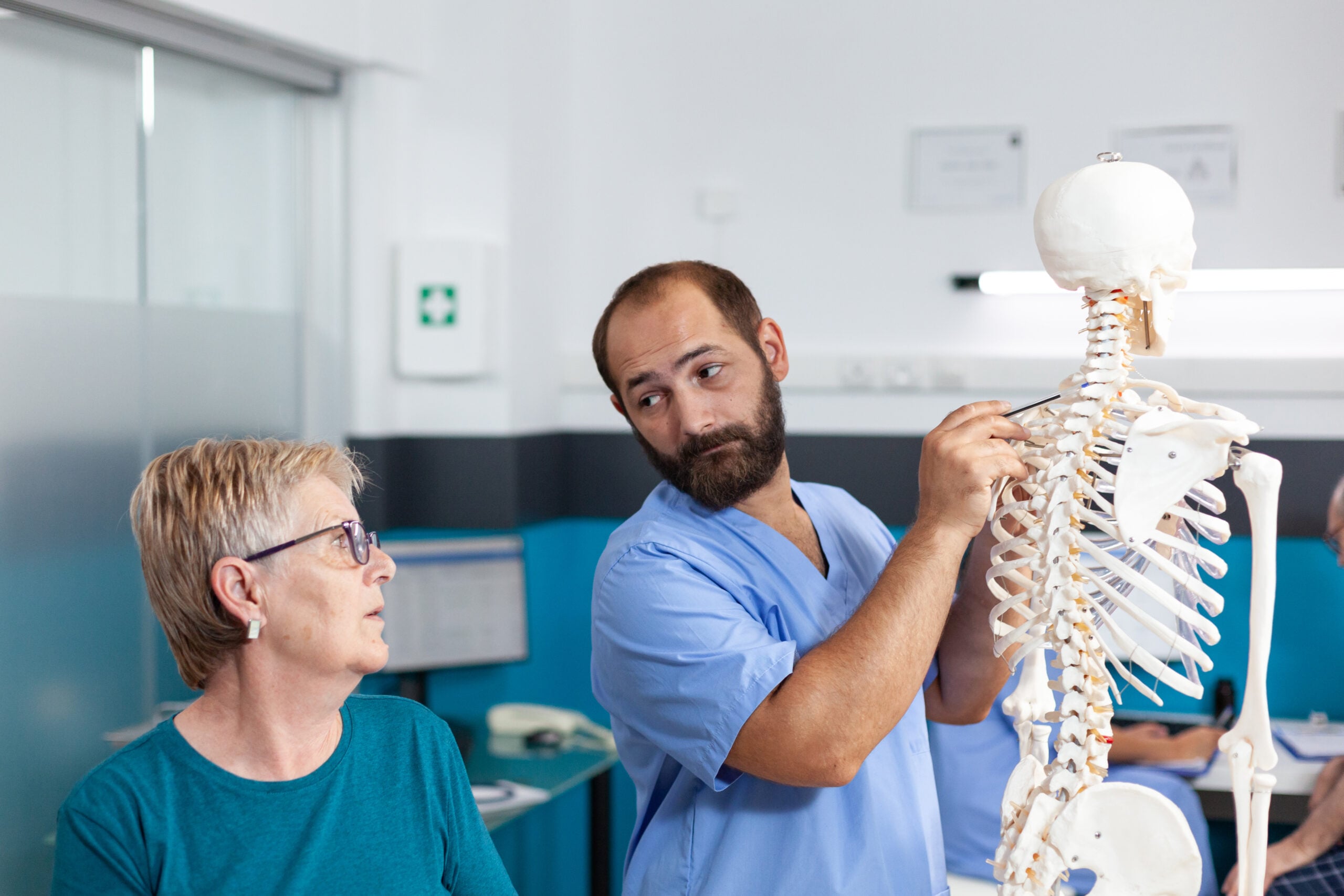 Woman and nurse looking at spinal cord on human skeleton for osteopathic examination. Chiropractor pointing at spine bones to explain diagnosis to retired patient for healthcare.