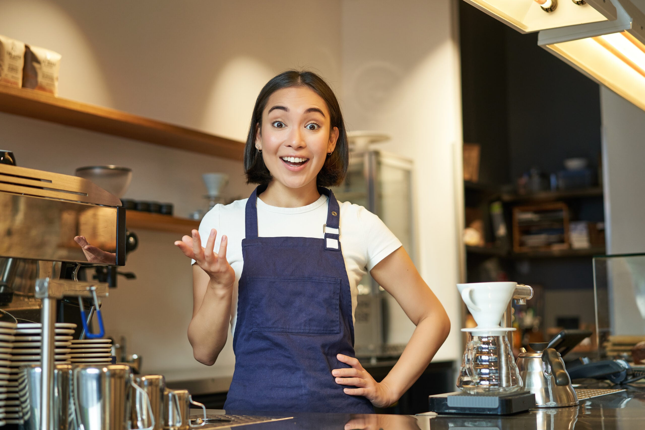 Portrait of girl barista in apron, looks surprised, points with hand and stares amazed at camera, works behind counter and brews coffee in highest paying teen jobs