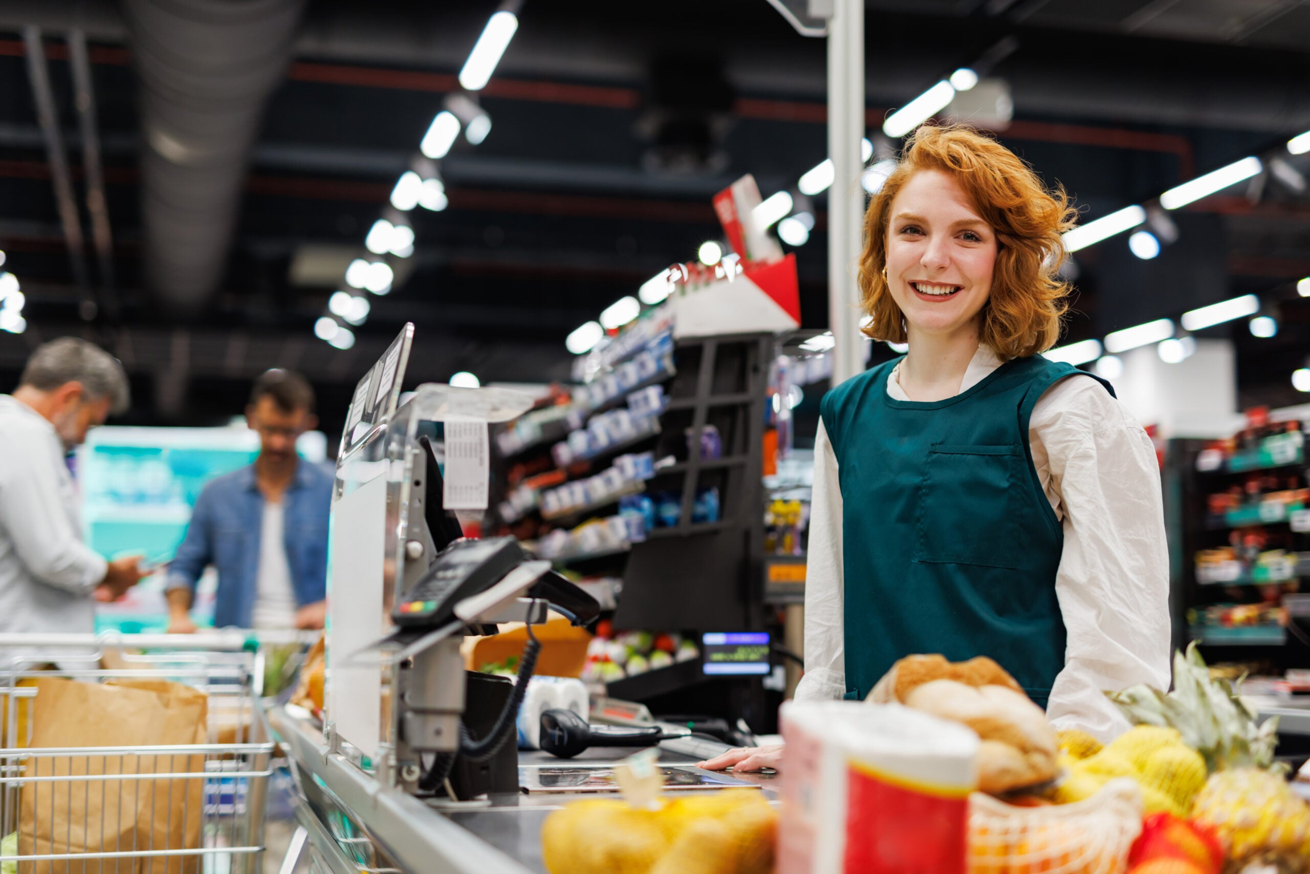 Redhead supermarket cashier wearing uniform is smiling while working at the checkout counter, scanning groceries for customers