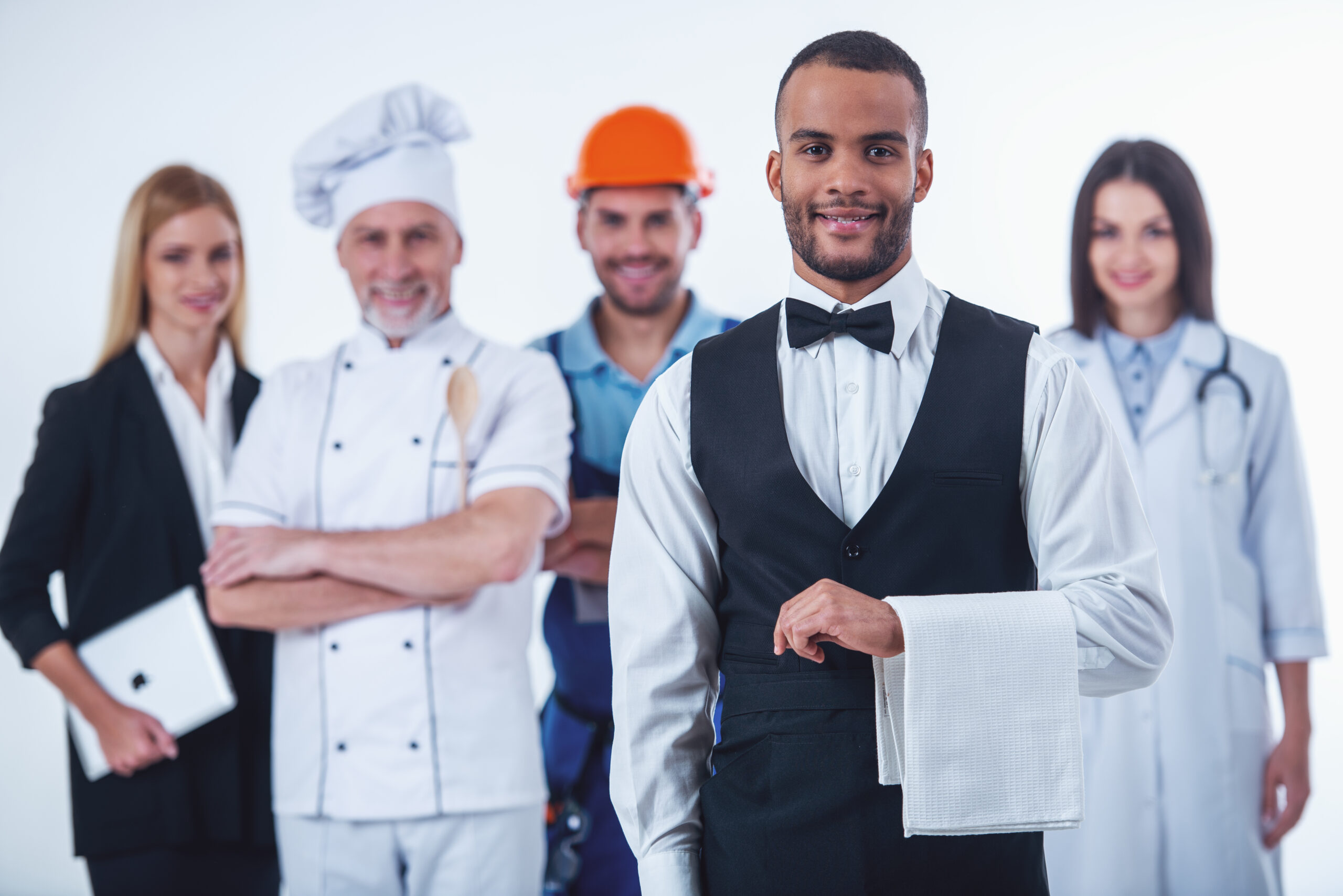 Beautiful people of different casual jobs in uniforms are looking at camera and smiling, isolated on white. Handsome Afro American waiter in the foreground