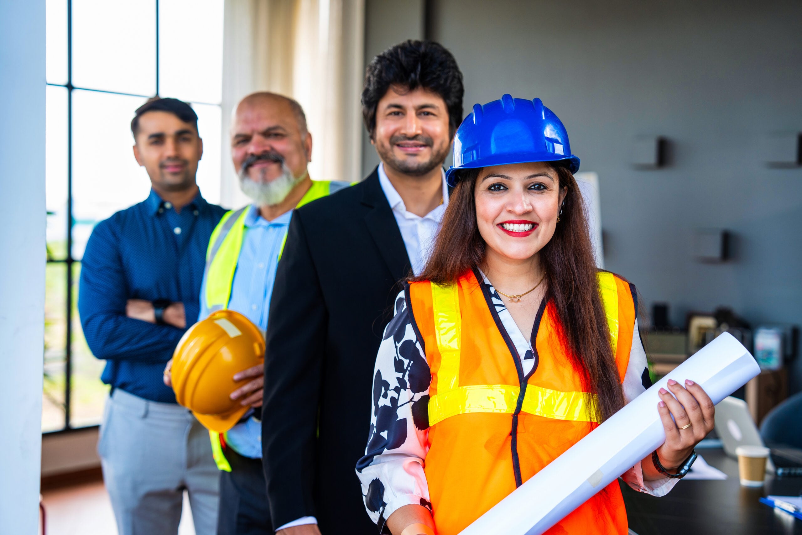 Indian Asian group of civil engineers and architects wearing fluorescent safety jackets, hard hats, holding rolled blueprints, standing confidently with folded arms, smiling, looking at camera