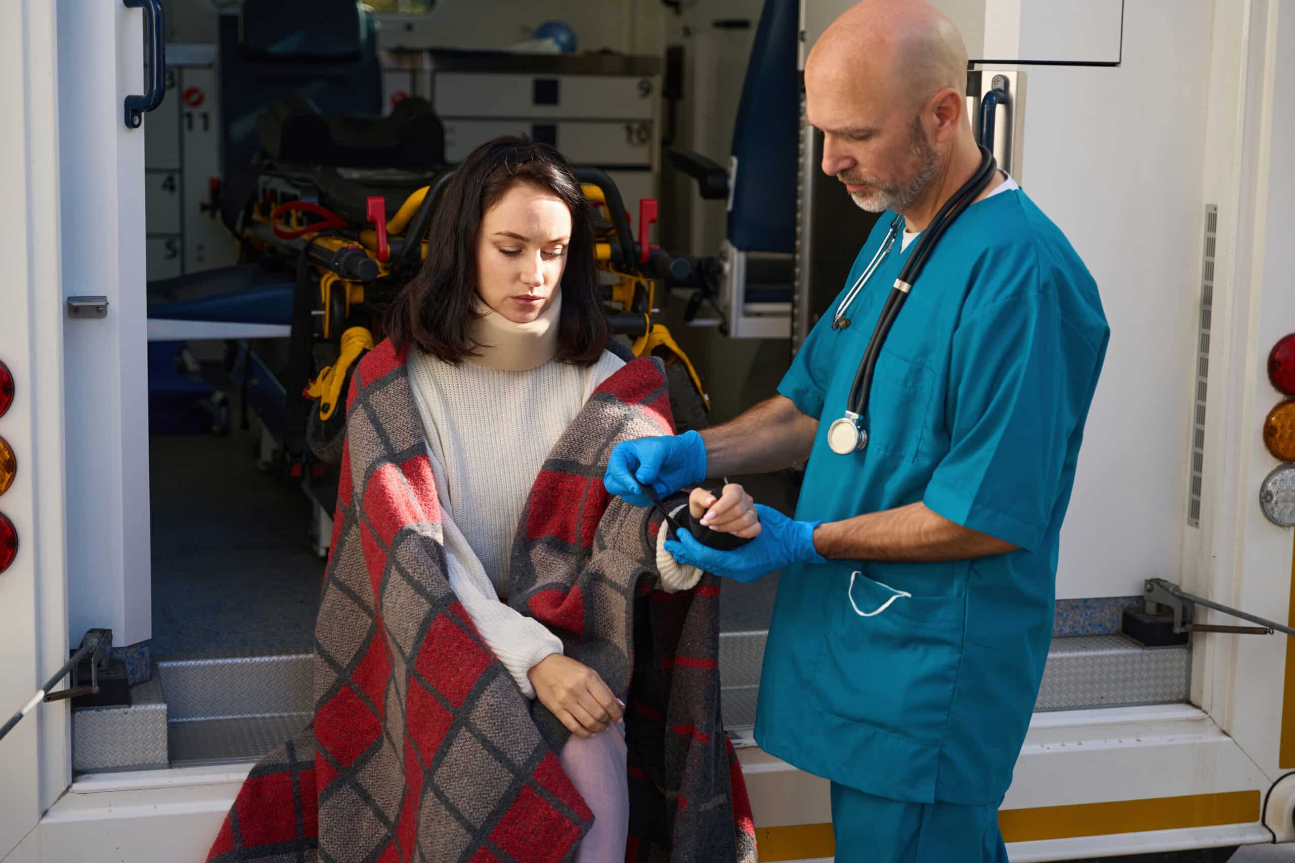 Sad patient sits wrapped in a blanket on the edge of an ambulance while a man fixes a fixing bandage on his wrist