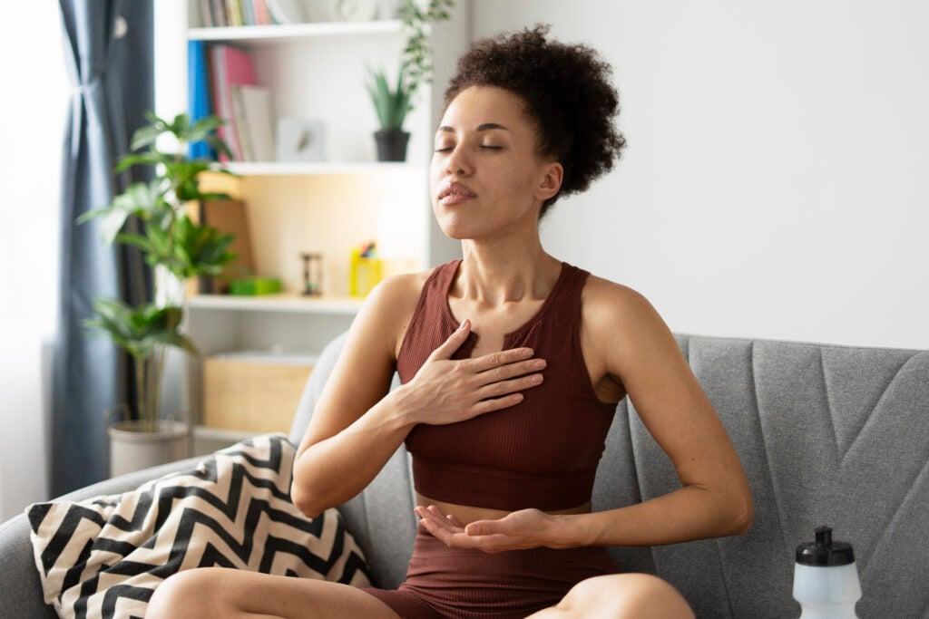 Serene young woman practicing mindfulness and deep breathing exercises, sitting in lotus position on her sofa, promoting relaxation and stress reduction through mindful breathing techniques
