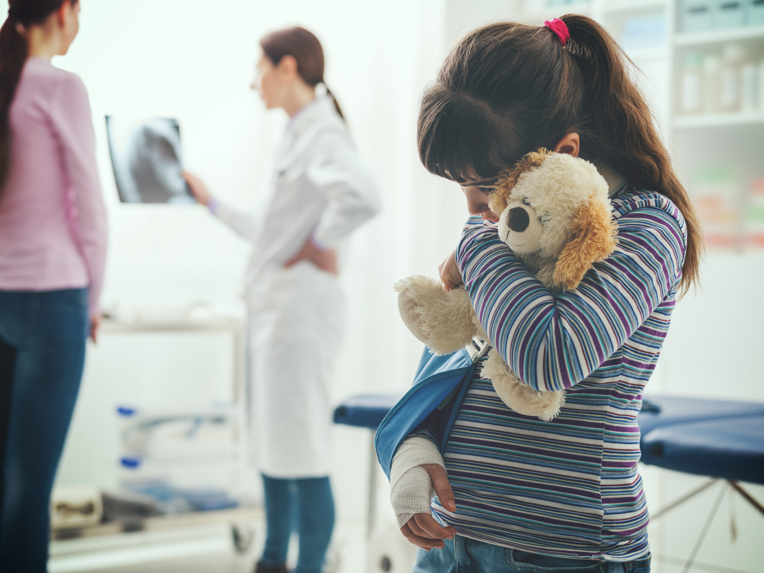 Scared cute girl with arm brace in the doctor's office, she is holding a teddy bear plushie, children and healthcare concept