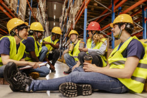 Diverse team of warehouse workers enjoying a coffee break, sitting on the floor amidst storage racks, fostering camaraderie and teamwork in a distribution center environment national safe work month