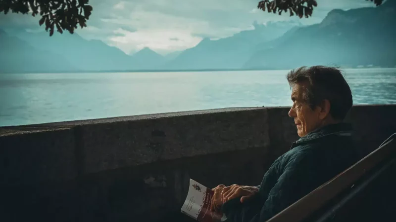 A warmly dressed old man sits, with book in hand, on a shady bench looking out over a great lake framed by huge mountains.