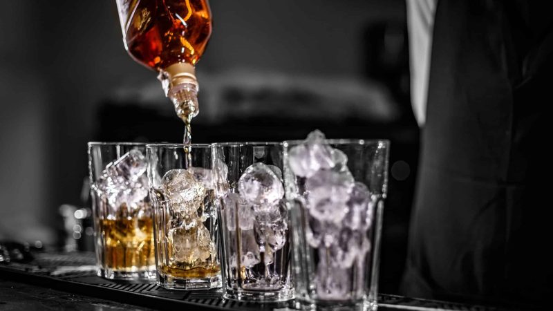 Barman pouring alcoholic drink into the glasses with ice cubes on the bar counter
