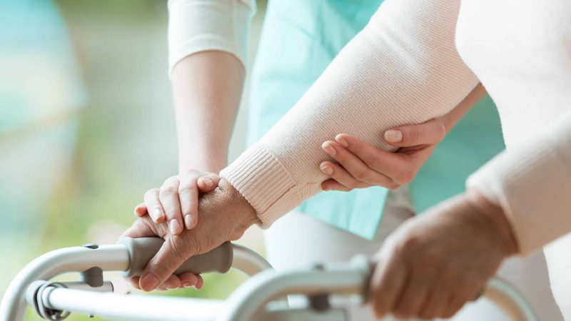 A disability support worker assists someone with a mobility aid.