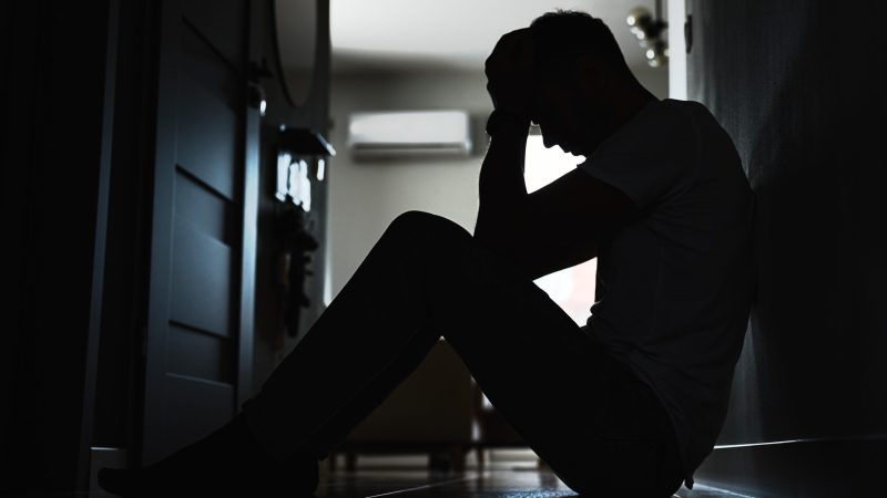 Depressed young man sitting on floor in dark room. Silhouette of young male suffering from loneliness and anxiety, covering his face with hands. Person with mental disorder antidepressants