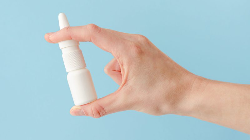 Female hand holding nasal spray on blue background. Seasonal allergy and common cold concept. neffy