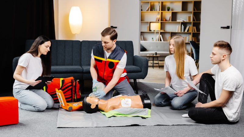 Young man instructor helping to make first aid heart compressions with dummy during emergency first aid training indoors.