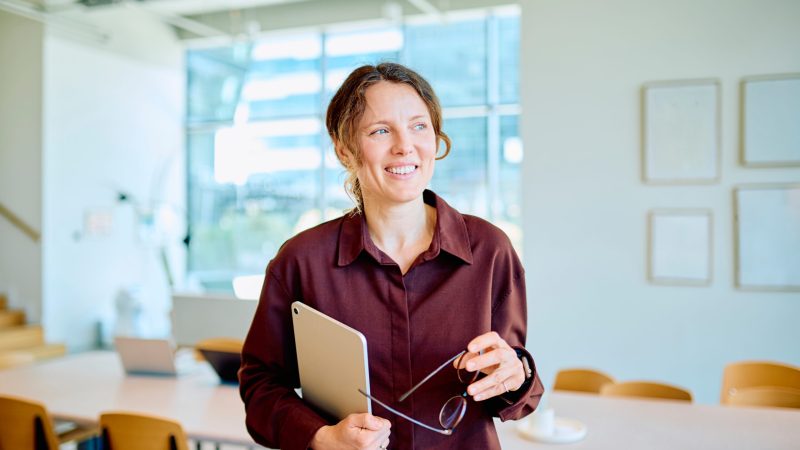 Professional woman smiling, holding a tablet and glasses, standing confidently in a bright, contemporary office environment Early Childhood Educators Day 2026