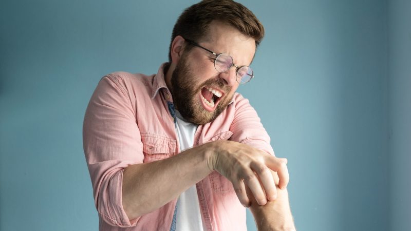 Unhappy, annoyed man scratches the itchy skin on arm, closes eyes and screams from the unpleasant sensation. Isolated, on a blue background. midge bites