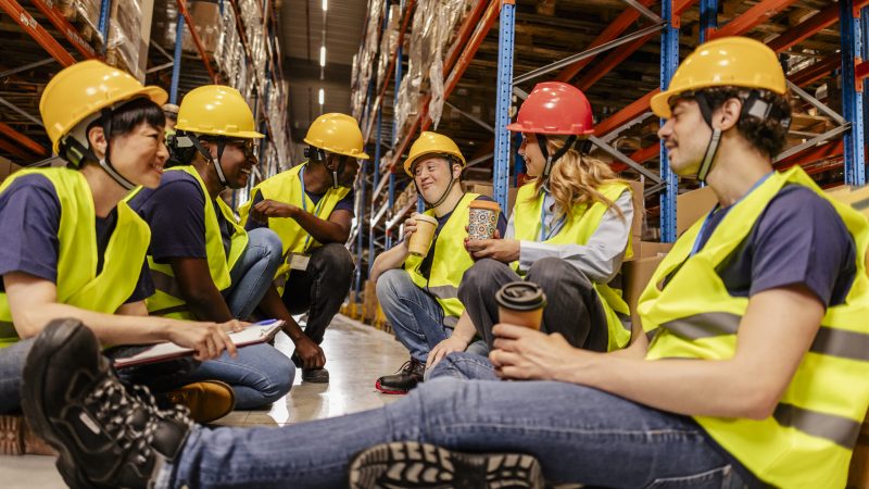 Diverse team of warehouse workers enjoying a coffee break, sitting on the floor amidst storage racks, fostering camaraderie and teamwork in a distribution center environment national safe work month