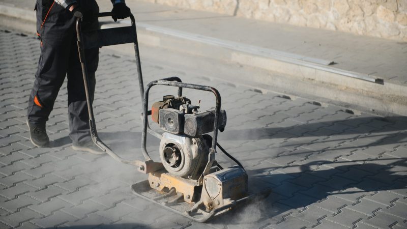 Male Worker using vibratory plate compactor to firm soil at worksite. builder. After laying of paving slabs, compacts paving stones with a gasoline vibratory compactor for subsequent. white card for paving jobs