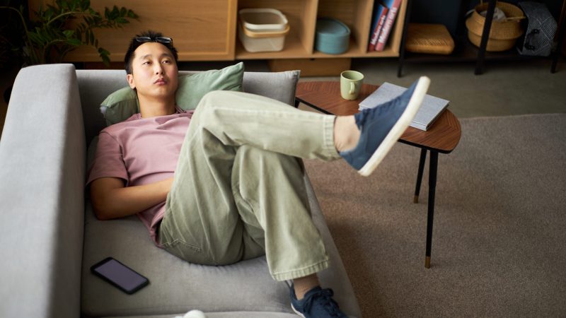 Young adult Asian man reclining on sofa with legs crossed, gazing upward with thoughtful expression, smartphone and headphones nearby on table, relaxing in modern living room sedentary lifestyle