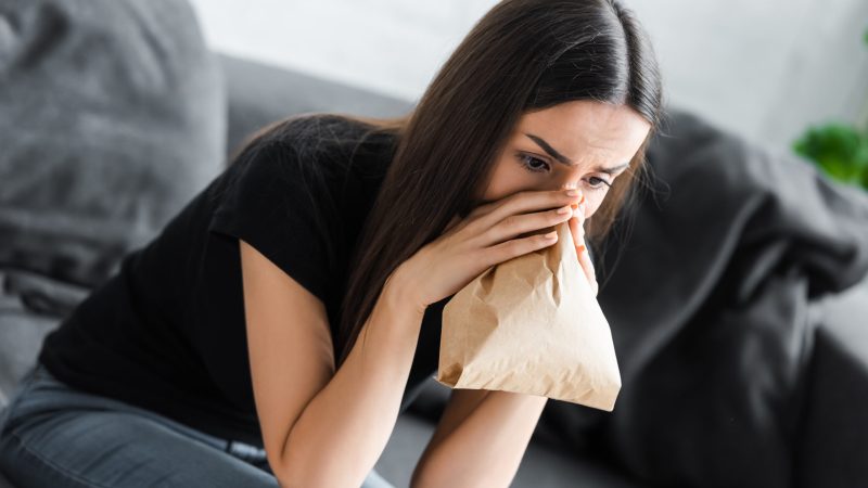 young woman hyperventilation into paper bag while suffering from panic attack at home